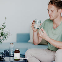 Man sitting on a couch in a light living room, holding a glass of water and a capsule, preparing to take a MAAX Boost supplement placed on the coffee table in front of him.